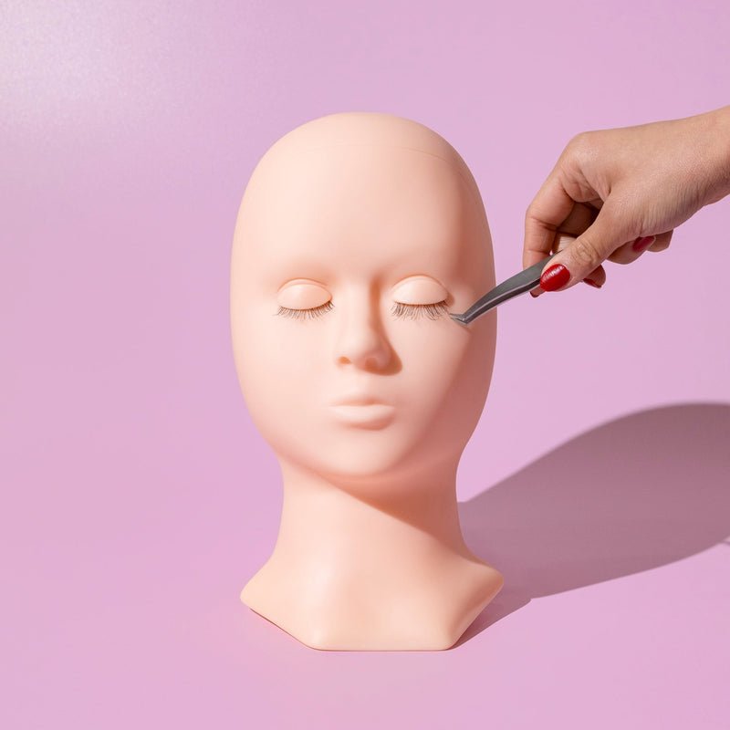 Lash training mannequin head with a hand using tweezers to apply eyelash extensions, set against a pink background.