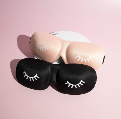 A pink and a black 3D contoured sleep mask with eyelash prints, placed on a soft pink surface.