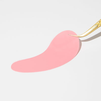 Curacoro gold tweezers holding a flat pink lash pad on a white background.