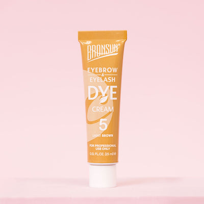 Light brown eyebrow and eyelash dye cream tube displayed on a pink background.