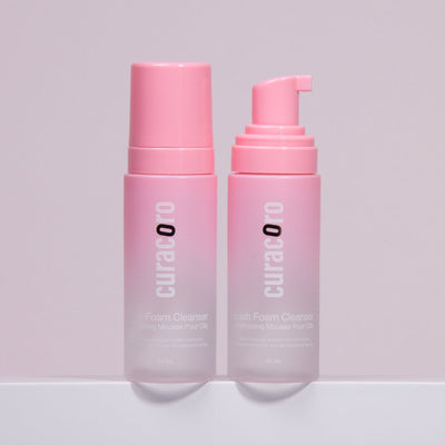 Two bottles of Curacoro Lash Foam Cleanser with pink pumps, one dispensing foam.