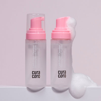 Two bottles of Curacoro Lash Foam Cleanser with pink pumps, one dispensing foam.