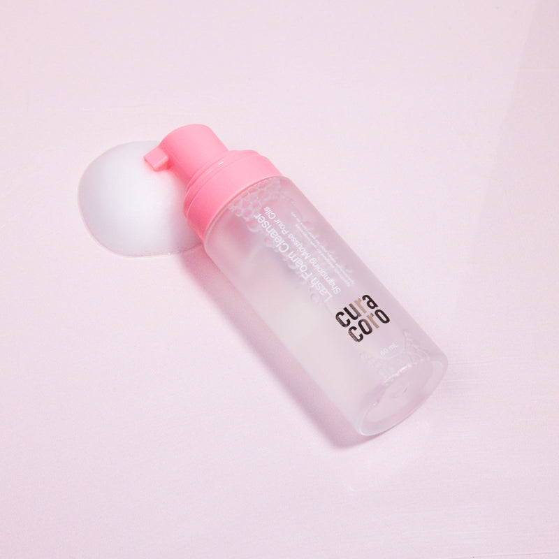 Curacoro Lash Foam Cleanser with a pink pump, lying on a light pink surface, surrounded by foam.