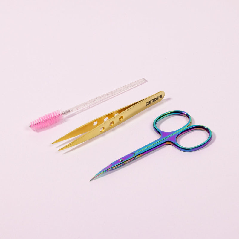 Curacoro lash extension tools set with pink mascara wand, gold precision tweezers, and iridescent scissors on soft pink background