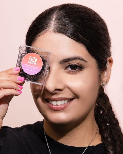 A smiling woman with braided hair holding a Curacoro Promade Loose Fans case near her eye against a soft pink background.