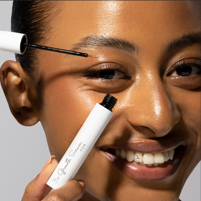 A smiling woman applying lash growth serum with an applicator to their upper lash line.