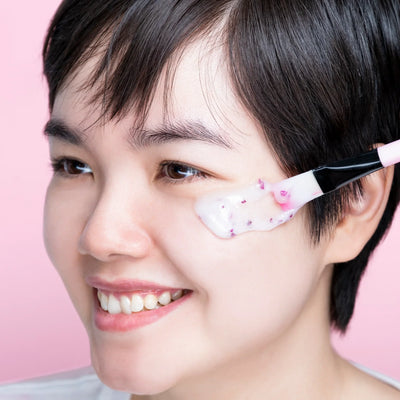 A smiling woman applying a rose hydro gel mask to her cheek against a pink background.