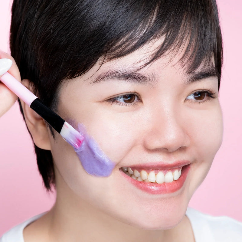 A smiling woman applying a Lavender hydro gel mask to her cheek against a pink background.