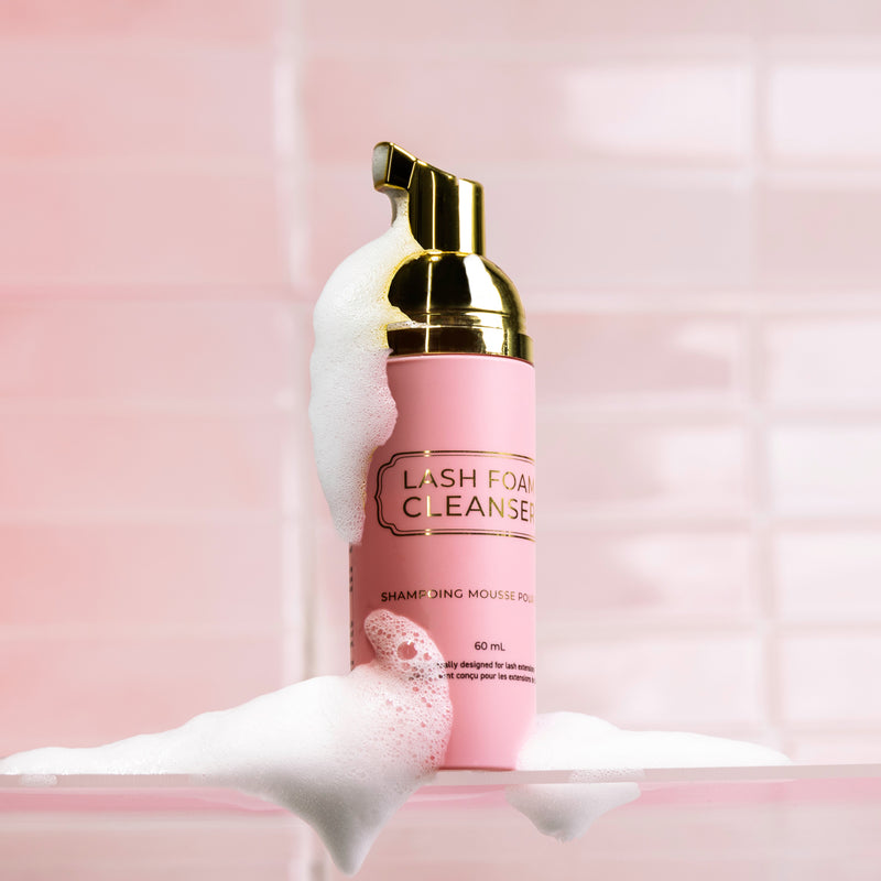 Lash Foam Cleanser bottle surrounded by white foam, set against a glossy pink tiled background.