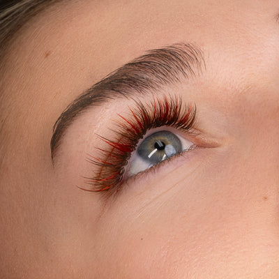 Close-up of a model's eye featuring wispy eyelash extensions in a mix of red and brown tones, showcasing a bold, vibrant look.