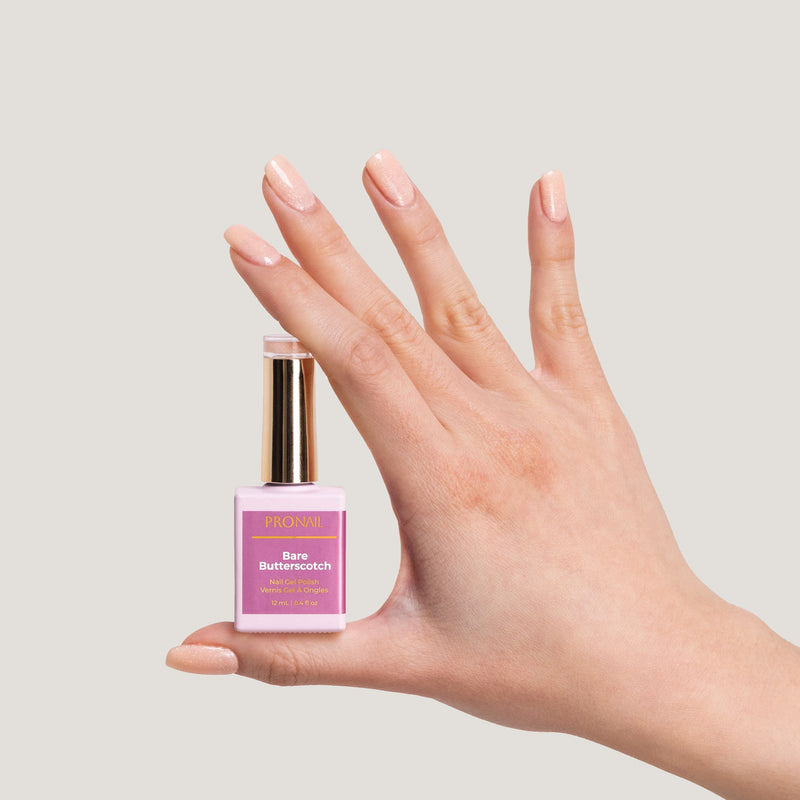 A hand with glittery Bare-butterscotch nails holding a nail polish bottle with a gold cap against a light background.