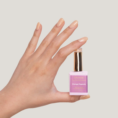 A hand with Orange-Popsicle-colored nails holding a nail polish bottle with a gold cap against a light background.