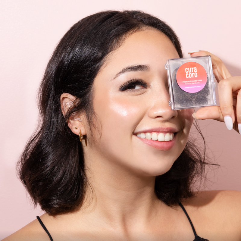 Smiling woman holding Curacoro promade loose fans case in front of her eye on a pink background.