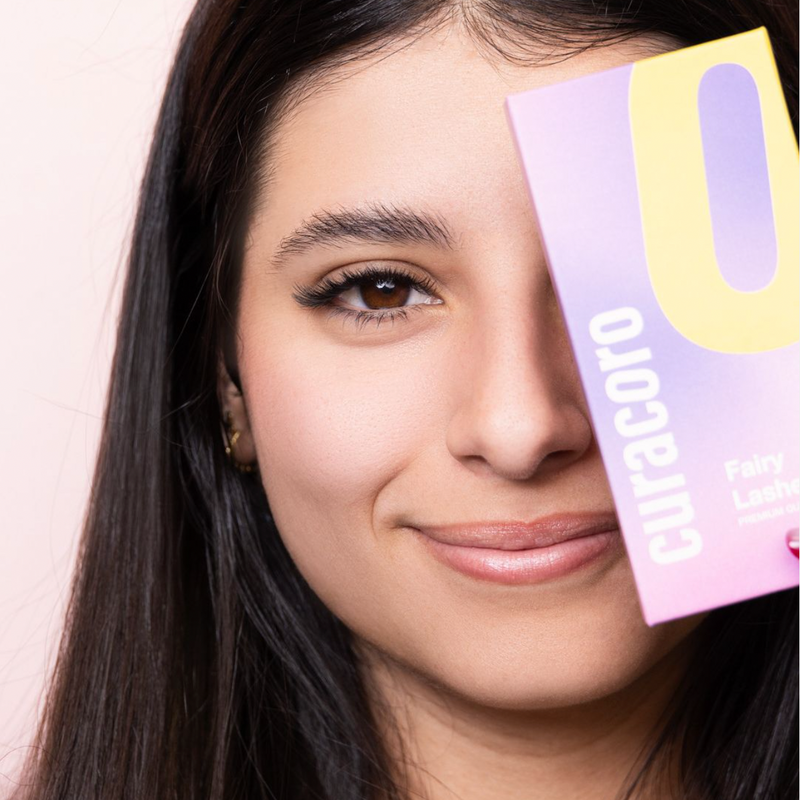 A close-up portrait of a smiling woman holding a Curacoro Fairy Lashes box, partially covering her face, against a light pink background.