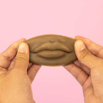 Hands holding a dark 3D Silicone Lips. against a pink background.