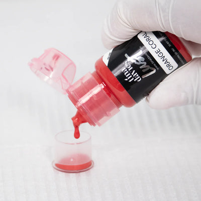 Gloved hand pouring Tina Davies Lust lip pigment in "Orange Coral" into a small clear cap.