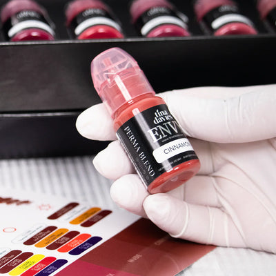 Gloved hand holding a Tina Davies Envy Cinnamon lip pigment bottle over a color chart, with pigment bottles in the background.