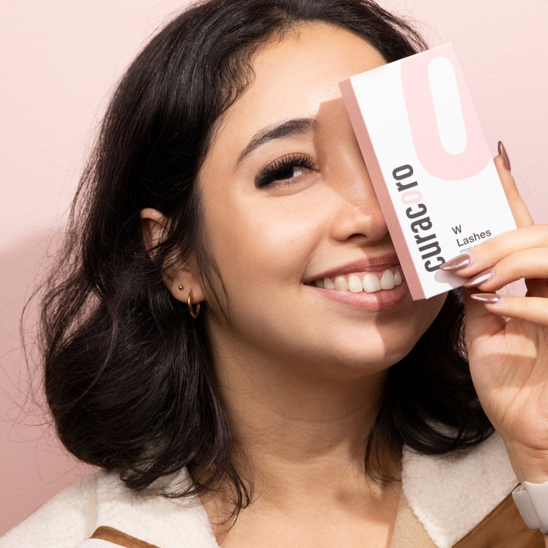 Smiling model holding Curacoro W Lashes packaging in front of her eye, showcasing her lashes look against a pink background.