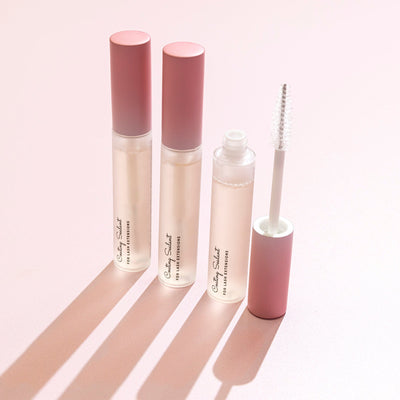 Three lash sealant tubes with pink ombre caps, one open with a clear applicator wand, arranged on a soft pink surface with shadows.