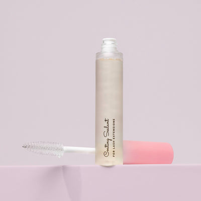 An open tube of lash sealant with a clear applicator wand, positioned upright on a soft pink surface against a light background.