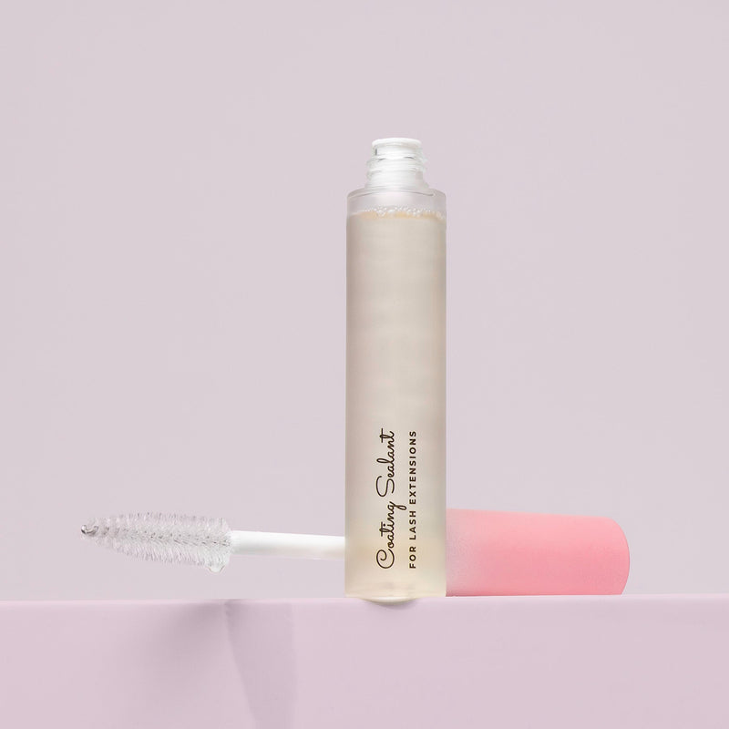 An open tube of lash sealant with a clear applicator wand, positioned upright on a soft pink surface against a light background.